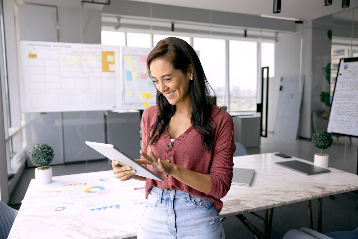 A woman looks at a tablet near a bulletin board and work table covered in notes.
