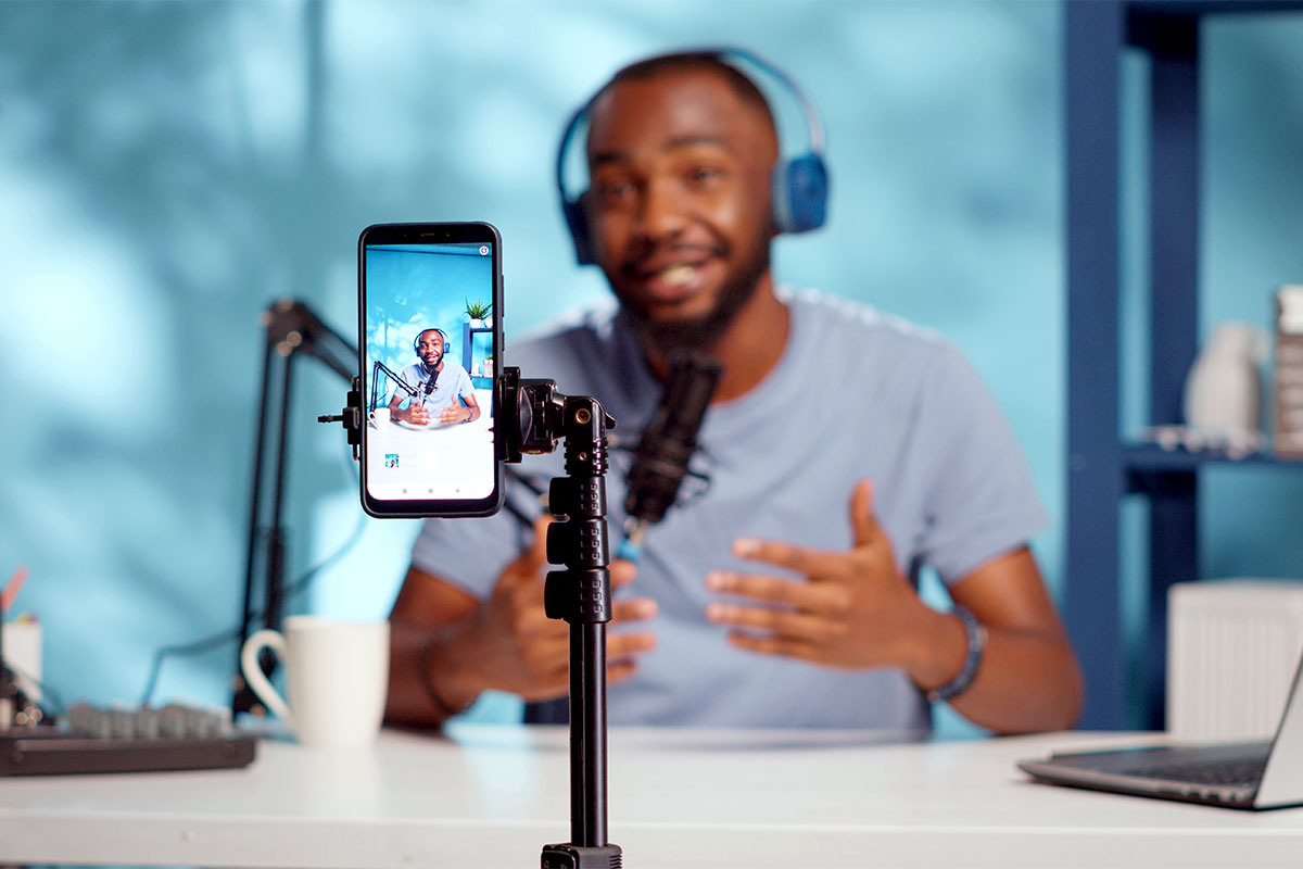 A man wearing headphones speaks into a microphone while being filmed with a cell phone on tripod.