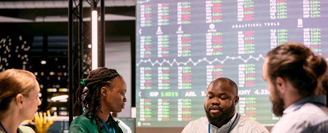 Four people sit around a conference table in discussion, with stock updates behind them.